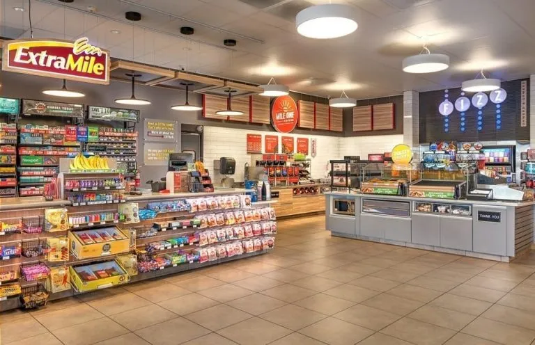 A convenience store interior showcases retail branding with shelves of snacks, drinks, and other items, plus a counter for prepared food and a self-serve beverage station in the background.