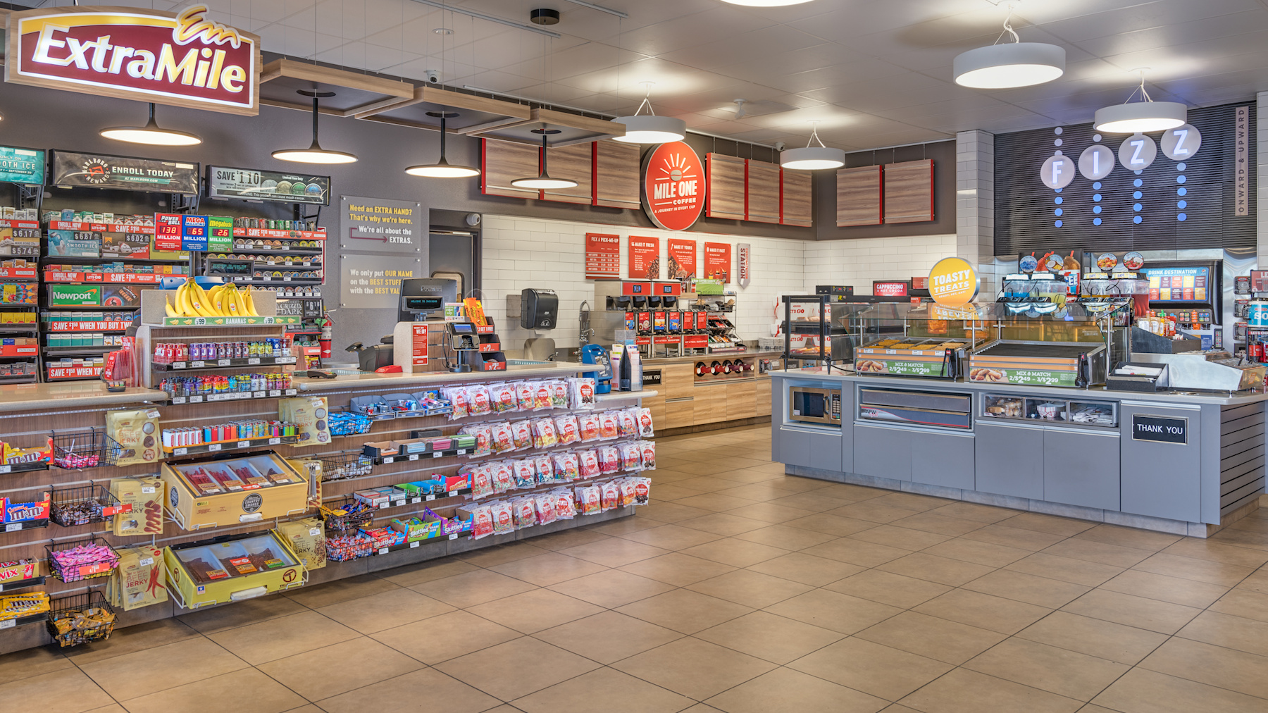 A convenience store interior showcases retail branding with shelves of snacks, drinks, and other items, plus a counter for prepared food and a self-serve beverage station in the background.